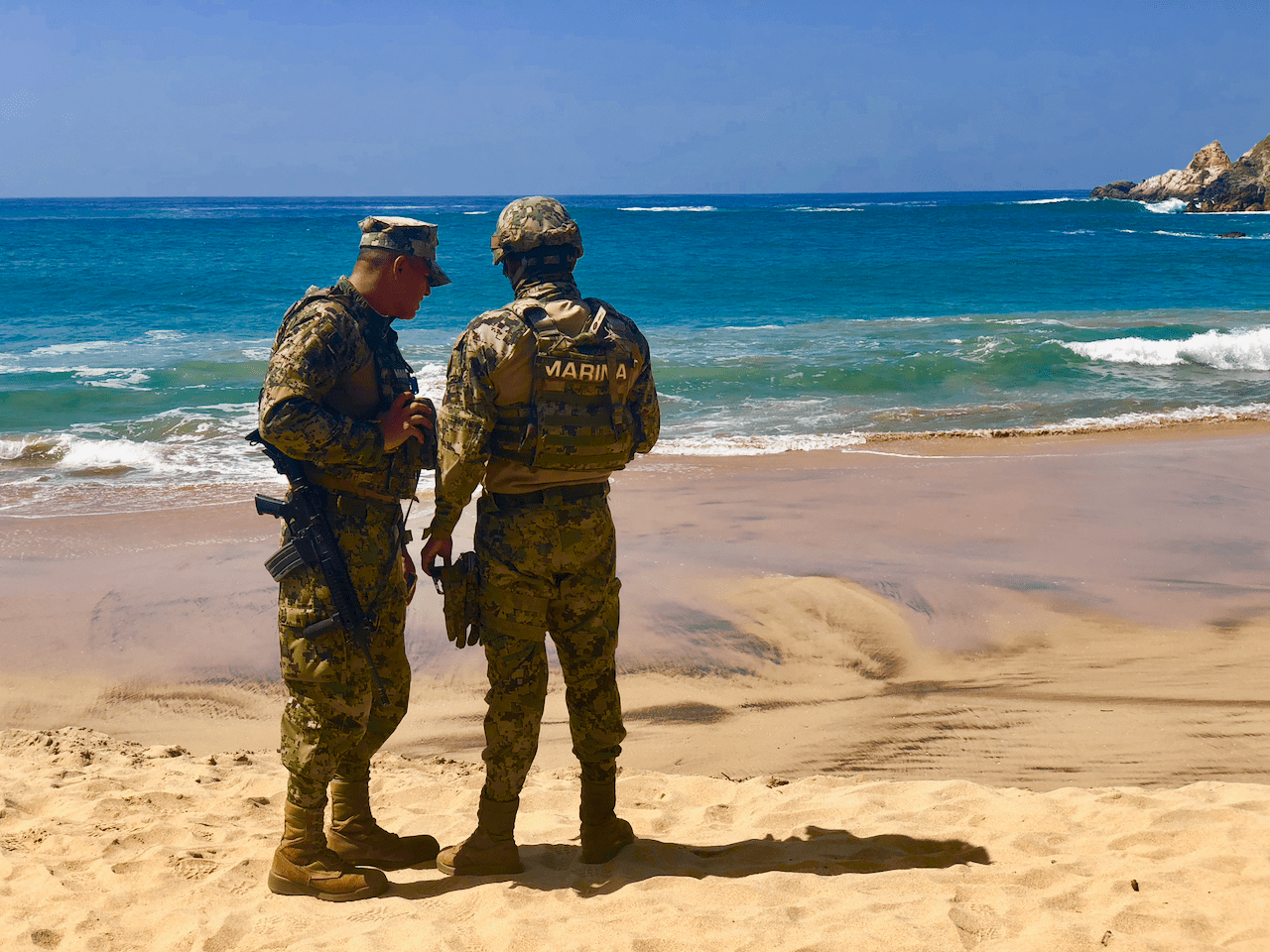 Two soldiers in camouflage uniforms standing on a beach looking out at turquoise waters